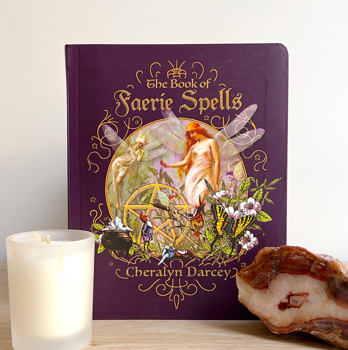 A beautifully bound copy of The Book of Faerie Spells by Cheralyn Darcey, featuring ornate gold detailing and artwork of faeries and nature on a rich purple cover, displayed on a wooden surface with a candle and crystal beside it. | Simply Esoteric