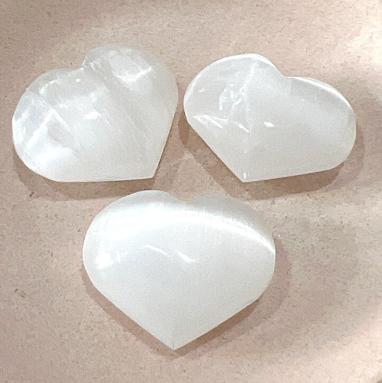 Three polished Selenite hearts, approximately 4cm in size, displaying their smooth, translucent surface on a soft pink background.