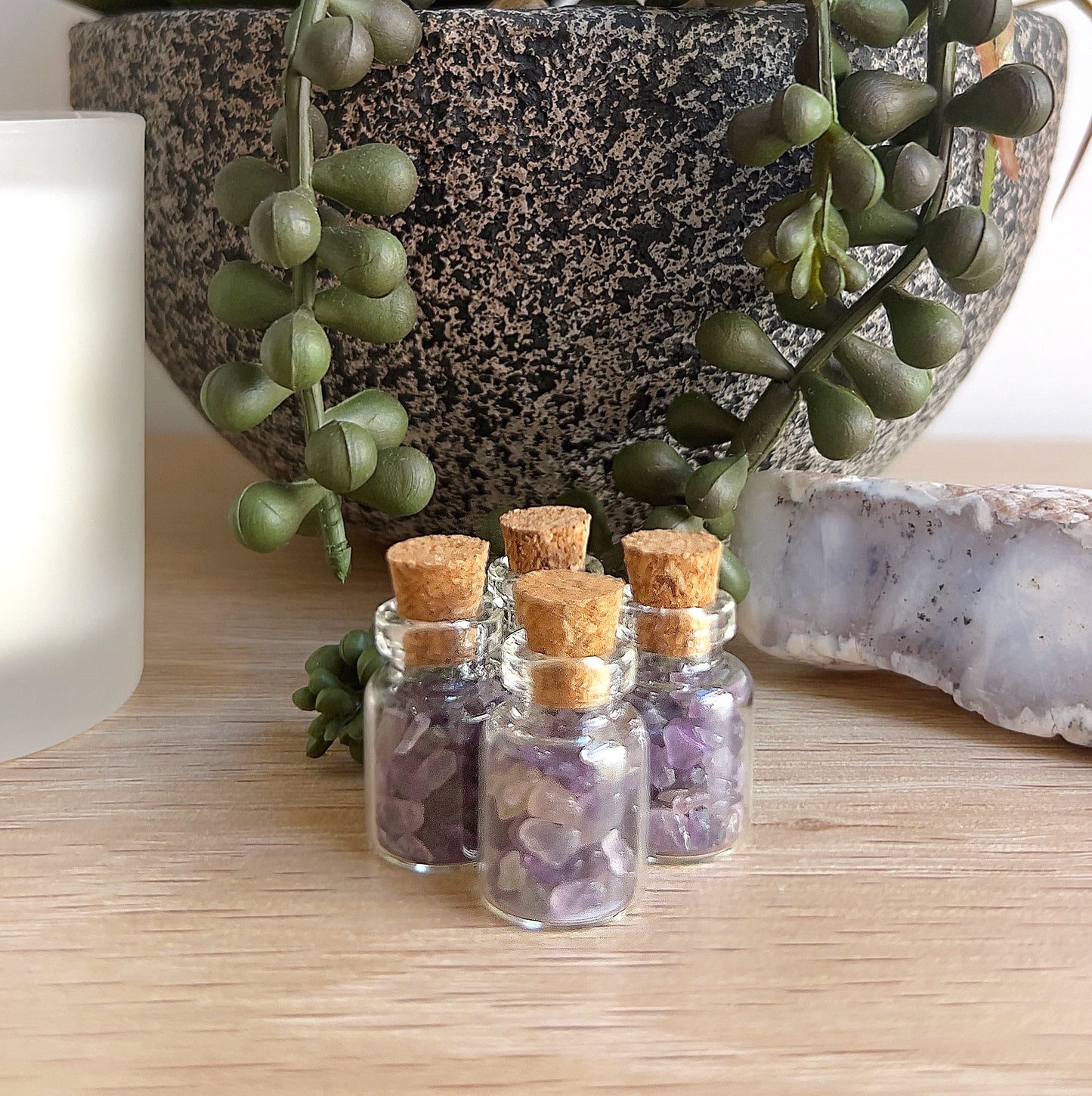 Four small glass bottles filled with delicate amethyst crystal chips, each sealed with a cork stopper, arranged neatly on a wooden surface alongside a succulent plant, white candle, and natural crystal, creating a tranquil and inviting setting. | Simply Esoteric