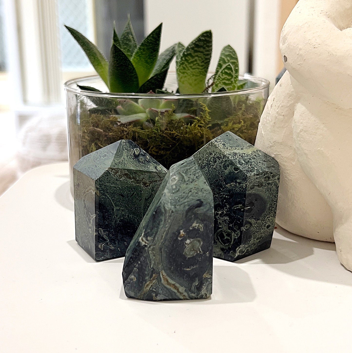 Three Kambaba Jasper Geoliths, approximately 5-6cm, featuring deep green and black swirling patterns, displayed on a light background with a plant in the background. | Simply Esoteric