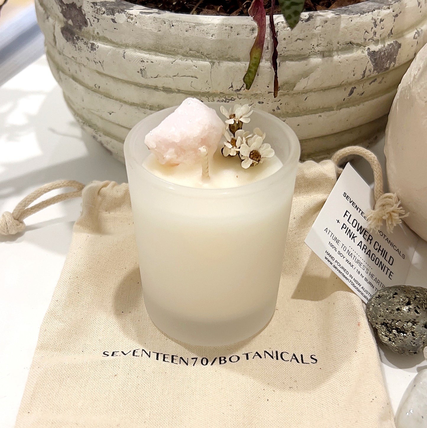 A crystal-infused soy wax candle from Seventeen70 Botanicals labeled "Flower Child + Pink Aragonite" sits on a white surface. The candle features a small pink aragonite crystal and scattered white flowers on top, creating a delicate and natural appearance. Accompanied by a label and a burlap drawstring bag, this product emphasizes a connection to nature with its eco-friendly and calming design. | Simply Esoteric