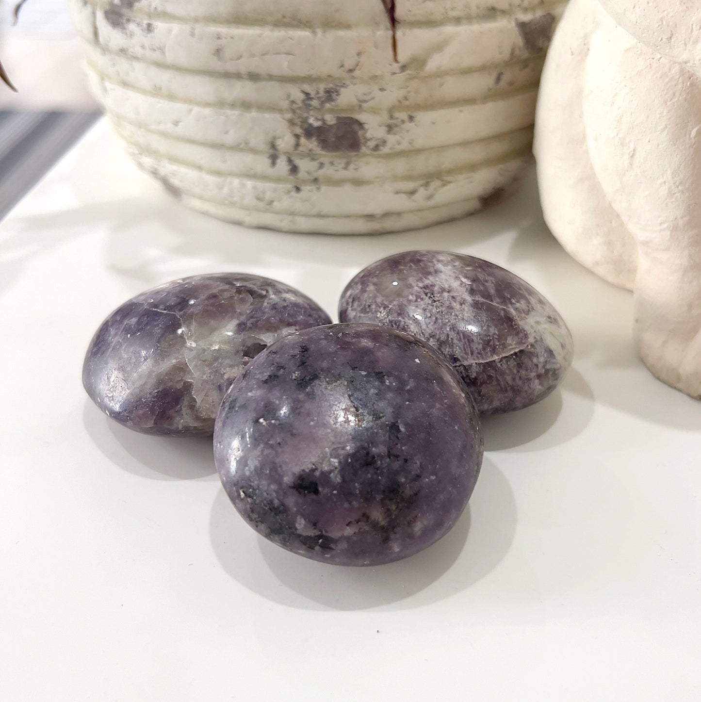 A trio of smooth, polished Lepidolite palm stones with a mix of deep purple and light lavender hues, placed on a white surface in front of a rustic, white ceramic pot with a plant. | Simply Esoteric