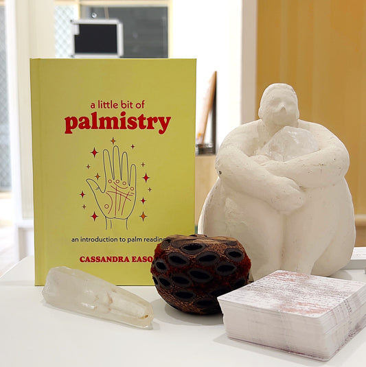 A photo of the book "A Little Bit of Palmistry" by Cassandra Eason displayed in a serene setting. The book cover features a simple design with a hand illustration showing palm lines. It is surrounded by decorative items, including a white crystal, a wooden seed pod, a stack of oracle cards, and a white statue, creating a peaceful and spiritual atmosphere.