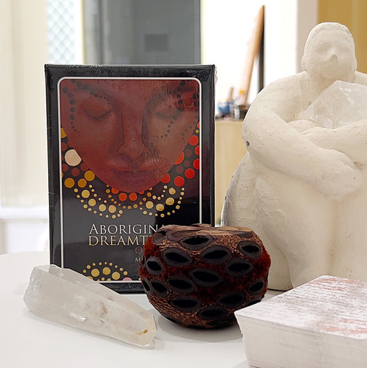 Aboriginal Dreamtime Oracle Cards by Mel Brown, displayed on a serene table setting with a white quartz crystal, an Australian native seed pod, and a hand-sculpted statue in the background, capturing the essence of spiritual wisdom and connection to Aboriginal culture