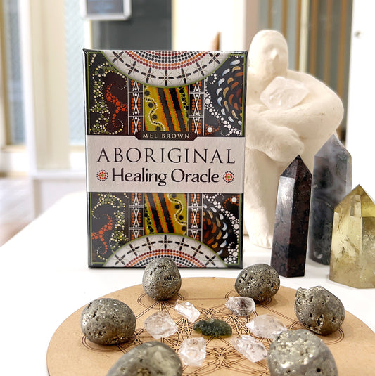 A set of Aboriginal Healing Oracle Cards by Mel Brown is displayed on a table. The box features colourful Aboriginal artwork with dot patterns. In front of the box, there are pyrite stones and clear quartz crystals arranged on a sacred geometry grid, adding a spiritual and healing vibe to the image. Crystal towers and a serene figurine are placed in the background, contributing to the peaceful and grounded energy of the scene.