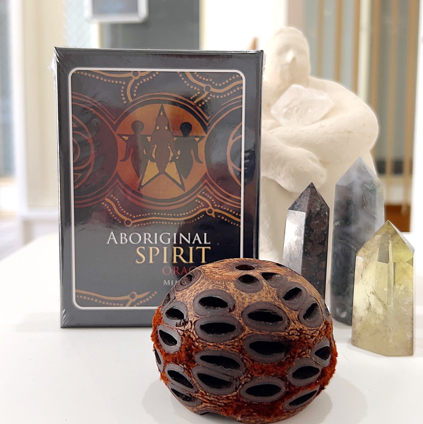 A deck of Aboriginal Spirit Oracle Cards by Mel Brown displayed on a white table. The box features Aboriginal artwork with a central star and figures in earthy tones. Surrounding the deck are several crystal towers and a decorative seed pod, creating a serene and spiritual atmosphere. The scene captures a connection between Indigenous wisdom and natural elements.
