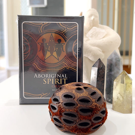 A deck of Aboriginal Spirit Oracle Cards by Mel Brown displayed on a white table. The box features Aboriginal artwork with a central star and figures in earthy tones. Surrounding the deck are several crystal towers and a decorative seed pod, creating a serene and spiritual atmosphere. The scene captures a connection between Indigenous wisdom and natural elements.