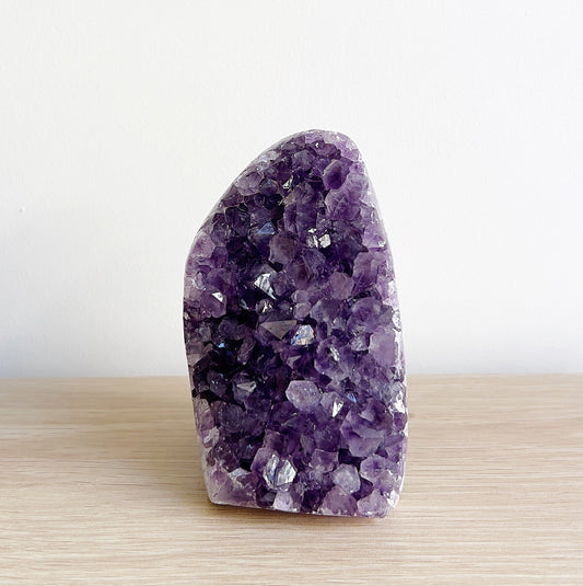 Close-up view of a sparkling amethyst crystal cluster, approximately 10cm tall, standing on a wooden surface against a minimalistic white background. | Simply Esoteric