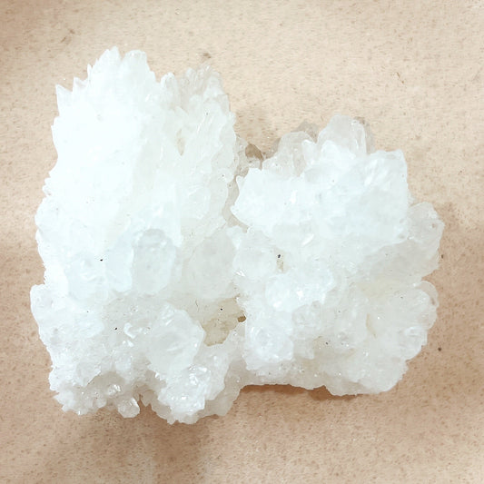 A close-up image of a natural white Aragonite crystal cluster, measuring approximately 6cm x 5.5cm x 3.5cm. The cluster features delicate, shimmering crystal formations, sitting on a neutral-toned surface. The intricate, sparkling design highlights the raw beauty of this grounding and stabilising stone.