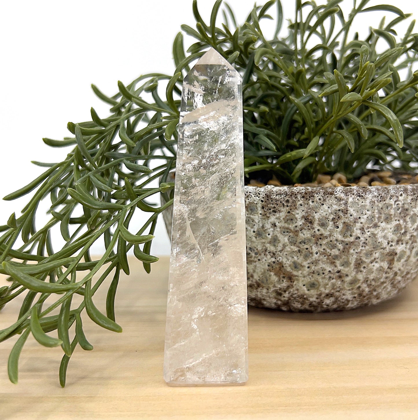 Clear Quartz obelisk crystal tower, 11cm tall, transparent with natural inclusions beside a textured stone pot and greenery — symbolising clarity, healing, and amplified energy.