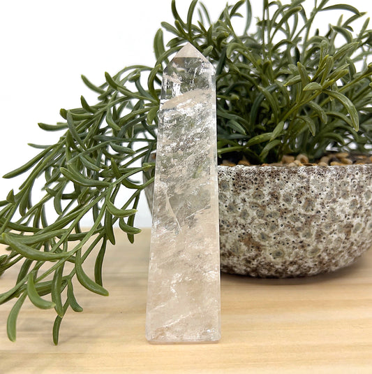 Clear Quartz obelisk crystal tower, 11cm tall, transparent with natural inclusions beside a textured stone pot and greenery — symbolising clarity, healing, and amplified energy.