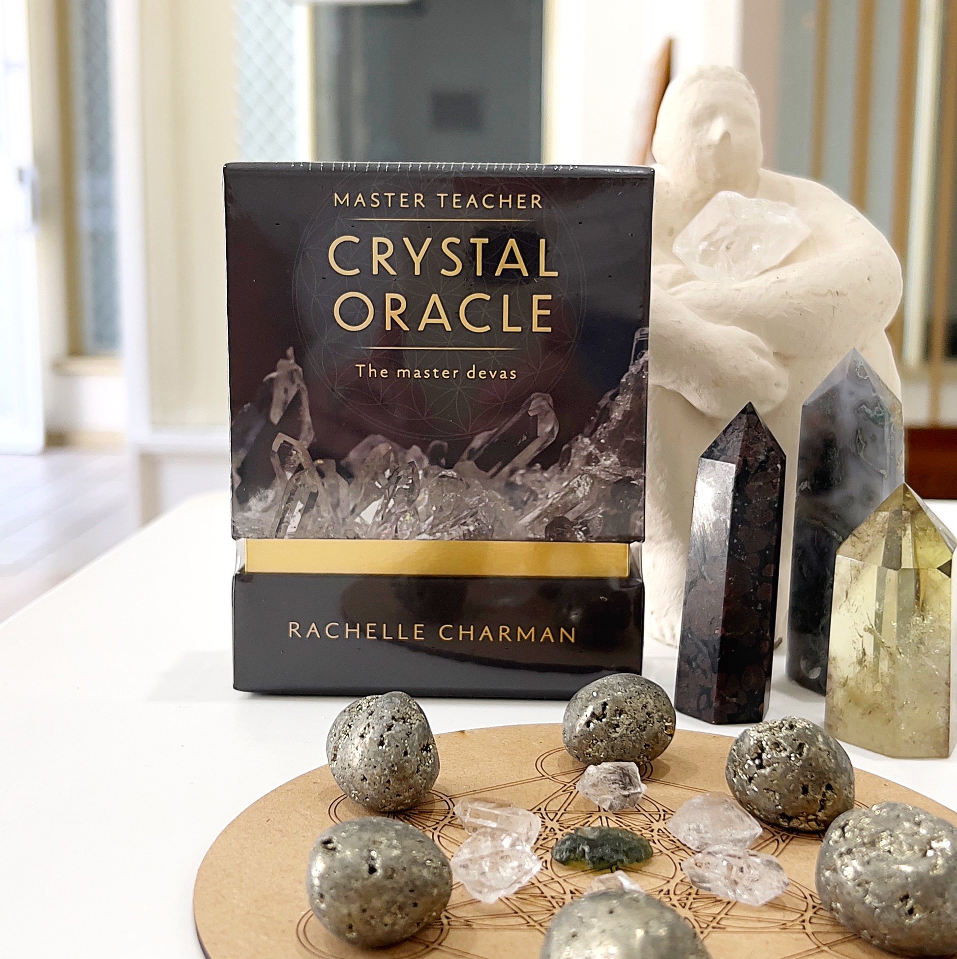 Crystal Oracle: The Master Devas by Rachelle Charman displayed on a table surrounded by pyrite stones, quartz crystals, and crystal towers. The box features a dark, elegant design with crystal imagery and golden accents.