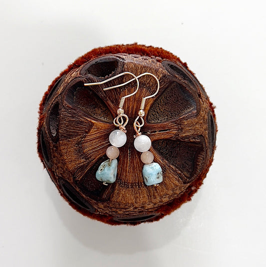 A pair of handmade earrings featuring Larimar, Rainbow Moonstone, and Peach Moonstone beads, elegantly arranged and hanging from gold hooks. The earrings are displayed on a rustic, natural brown seed pod, highlighting their earthy, spiritual essence and connection to Goddess energy.