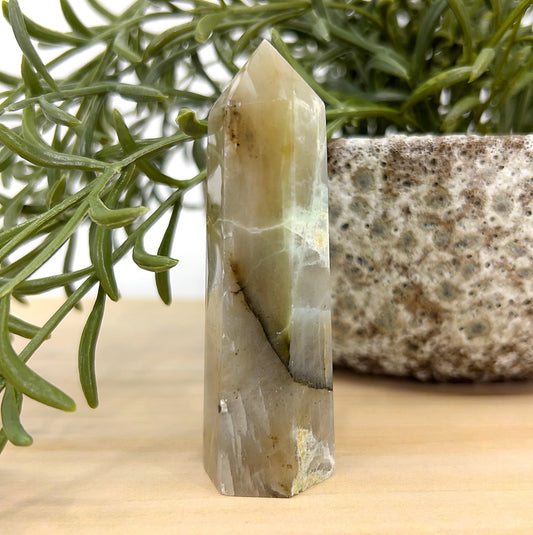 Green Moonstone crystal tower standing 8cm tall with soft green and cream hues, displayed beside a textured stone pot and green foliage — symbolising emotional balance, intuition, and heart healing.