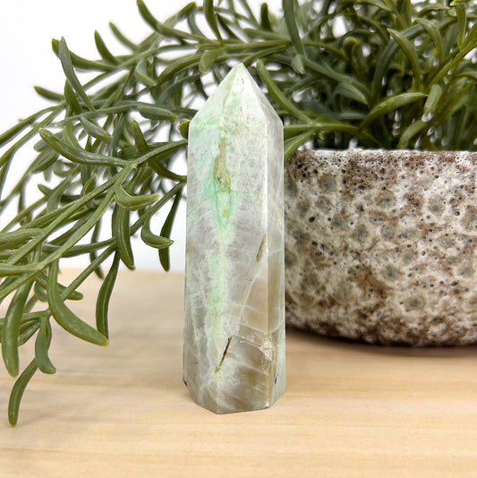 Green Moonstone crystal tower standing 8cm tall with soft green and cream hues, displayed beside a textured stone pot and green foliage — symbolising emotional balance, intuition, and heart healing.