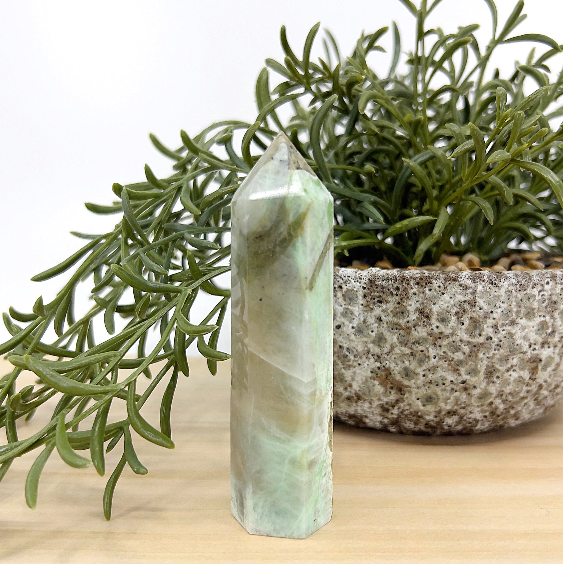 Green Moonstone crystal tower standing 10cm tall with soft green and cream hues, displayed beside a textured stone pot and green foliage — symbolising emotional balance, intuition, and heart healing.