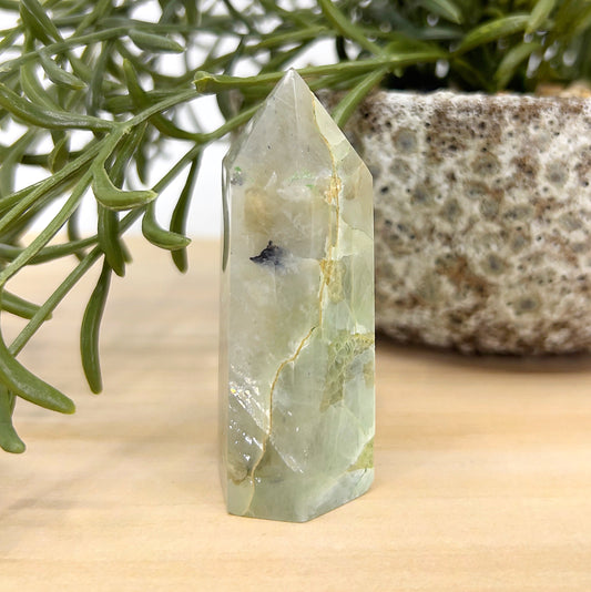 Green Moonstone crystal tower standing 7cm tall with soft green and cream hues, displayed beside a textured stone pot and green foliage — symbolising emotional balance, intuition, and heart healing.