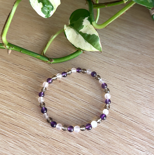 Beaded bracelet with Amethyst, Rose Quartz, and Smokey Quartz beads on a wooden surface with green leaves.