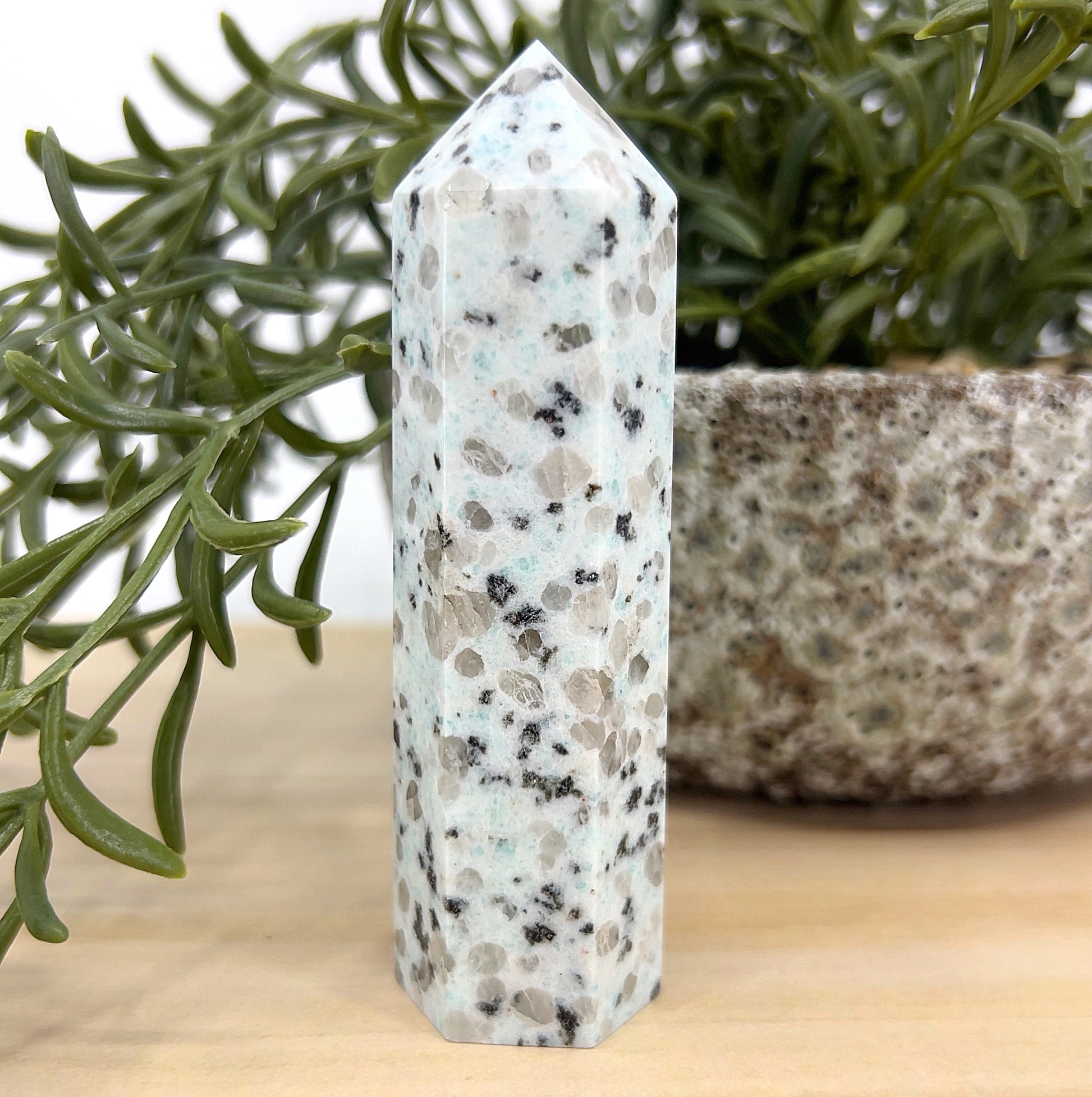 Kiwi Stone crystal tower approx. 10 cm tall with mint-green tones and black tourmaline speckles, displayed beside a green plant and stone pot – a healing crystal for calm energy and emotional balance.