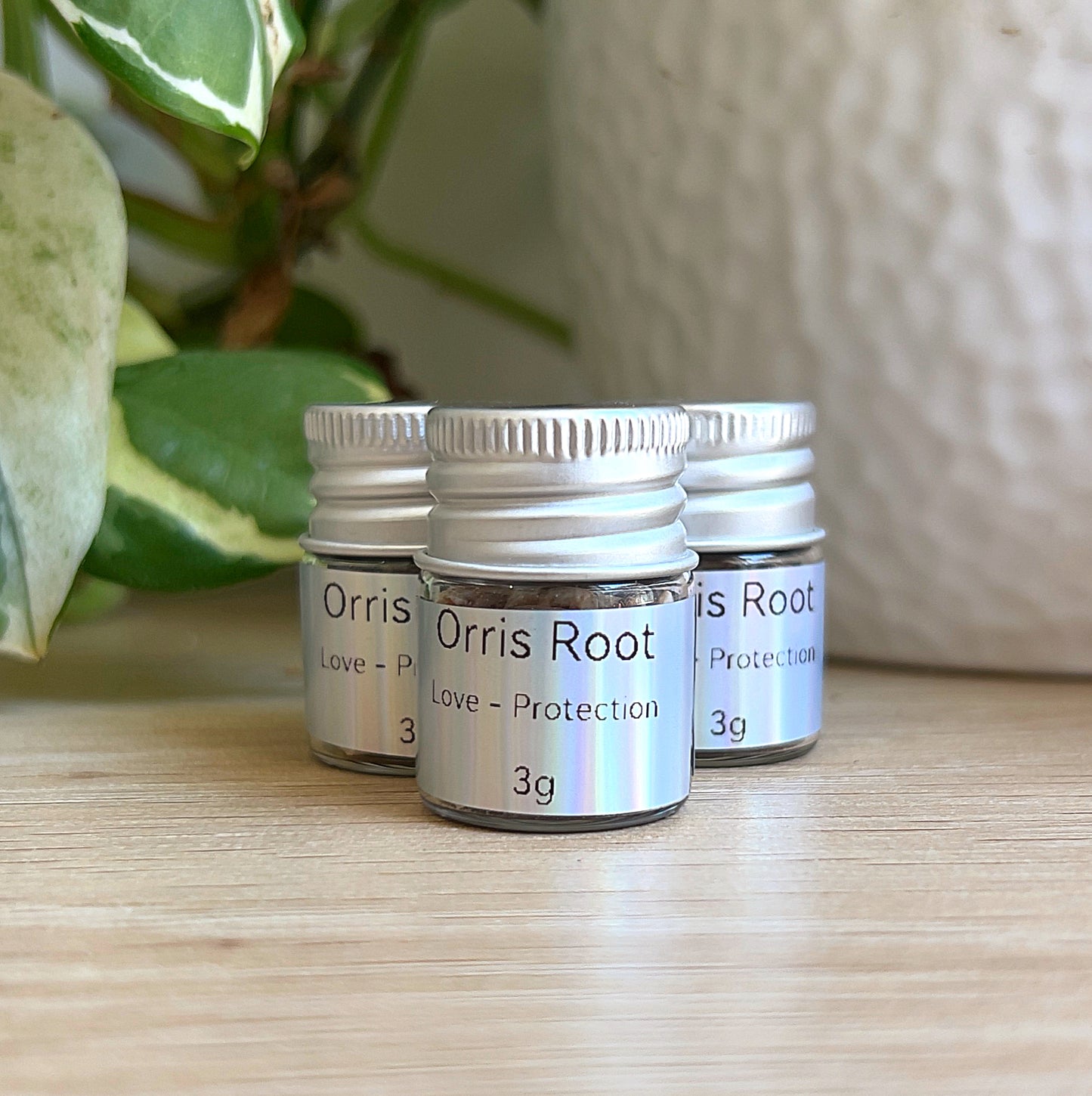 Small jars of orris root (3 g) with silver lids and labels, grouped on a wooden surface beside green plant leaves and a white pot.