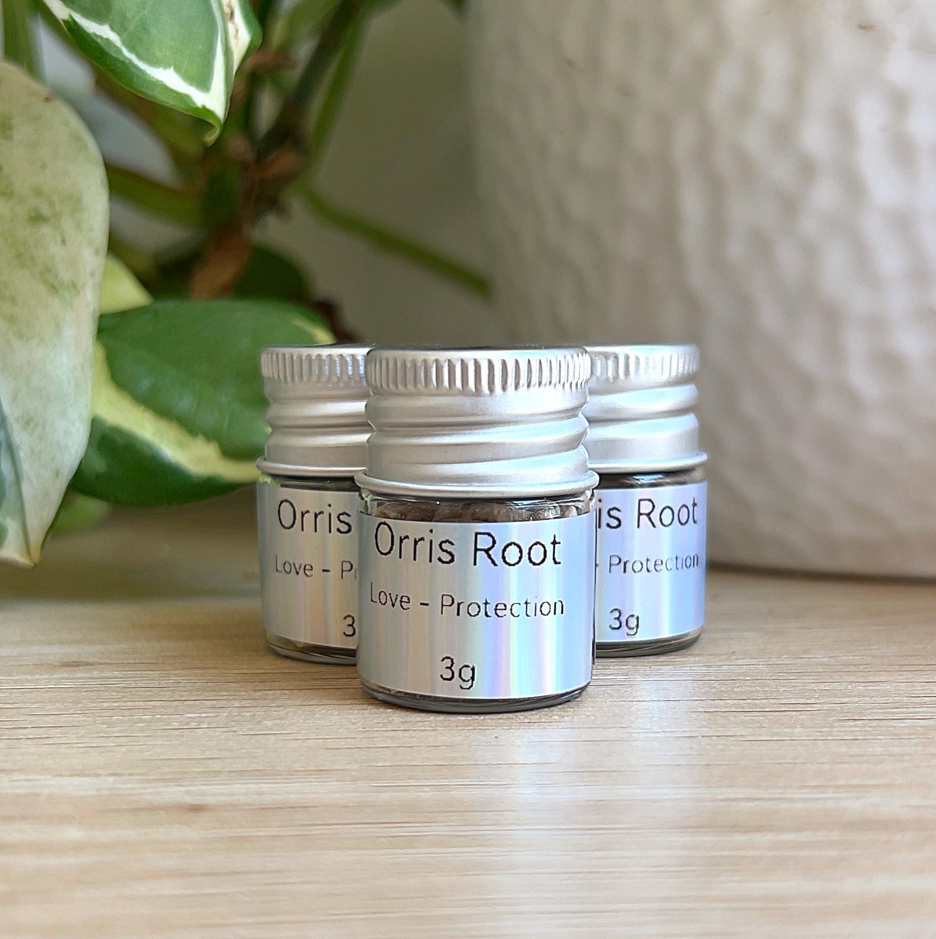 Small jars of orris root (3 g) with silver lids and labels, grouped on a wooden surface beside green plant leaves and a white pot.