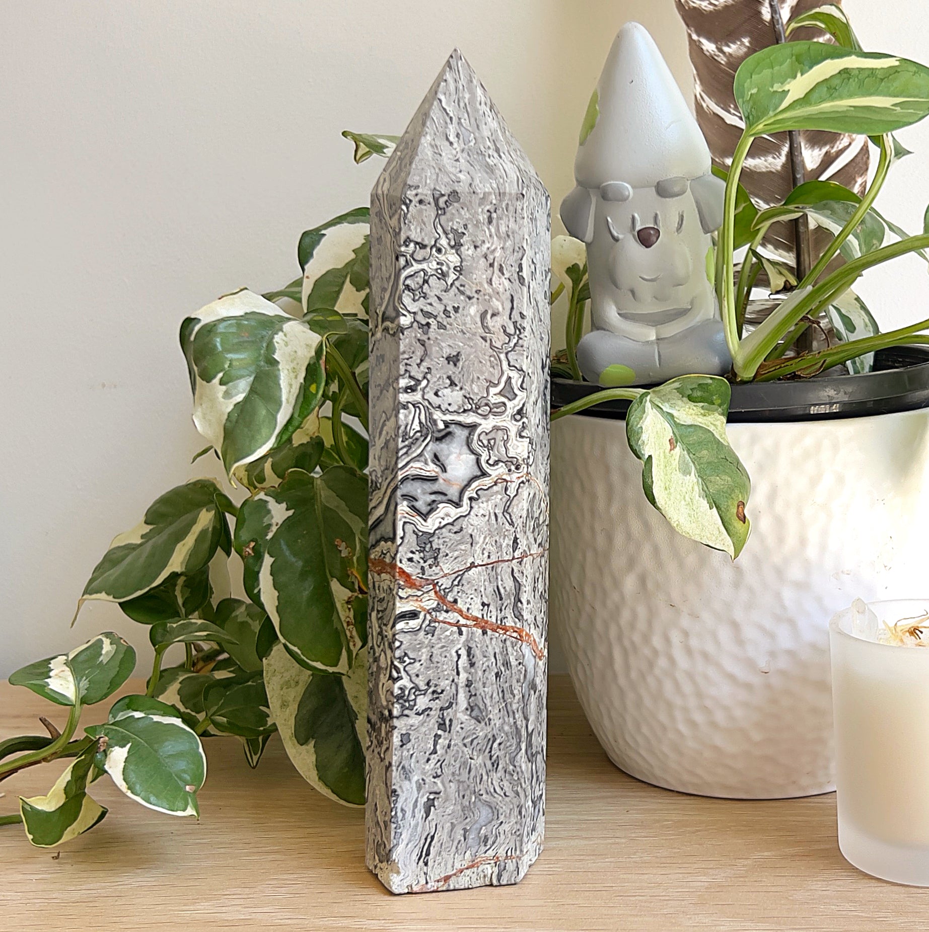 Polished Picasso stone tower (22 cm) with grey and black marbling and a rust-coloured vein, standing on a wooden surface beside variegated plant leaves and a white pot.