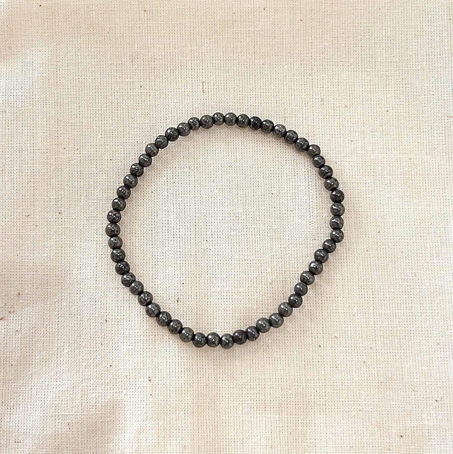 A Pyrite crystal beaded bracelet, featuring polished metallic grey Pyrite beads strung on a stretchy cord. The bracelet is laid flat on a neutral beige fabric background, showcasing the smooth and reflective surface of the beads. Ideal for promoting protection, abundance, and grounding.
