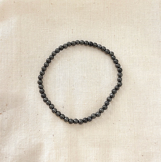 A Pyrite crystal beaded bracelet, featuring polished metallic grey Pyrite beads strung on a stretchy cord. The bracelet is laid flat on a neutral beige fabric background, showcasing the smooth and reflective surface of the beads. Ideal for promoting protection, abundance, and grounding.