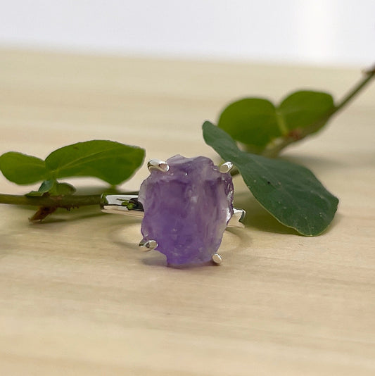 Ring with a raw Amethyst gemstone on a wooden surface with green leaves.