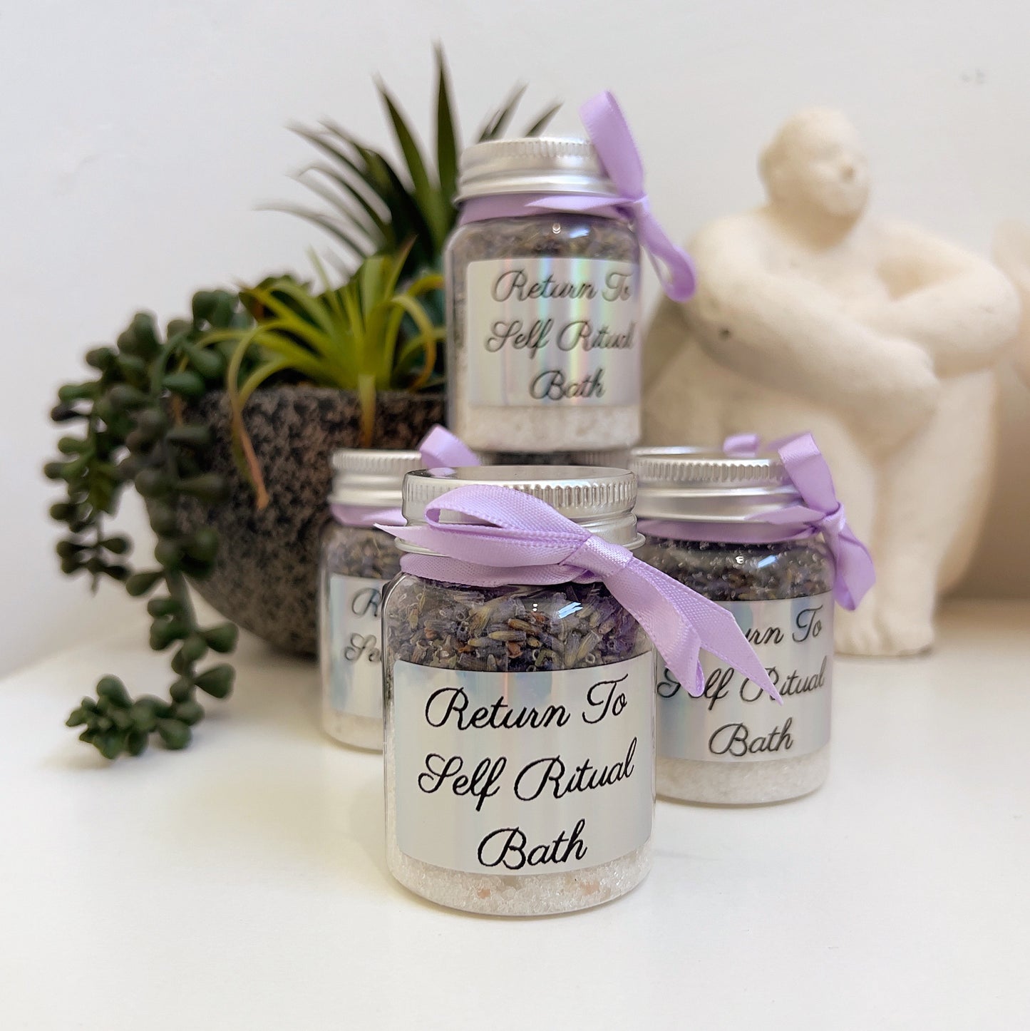 Jars labeled 'Return To Self Ritual Bath' with purple ribbons on a white surface with decorative items.