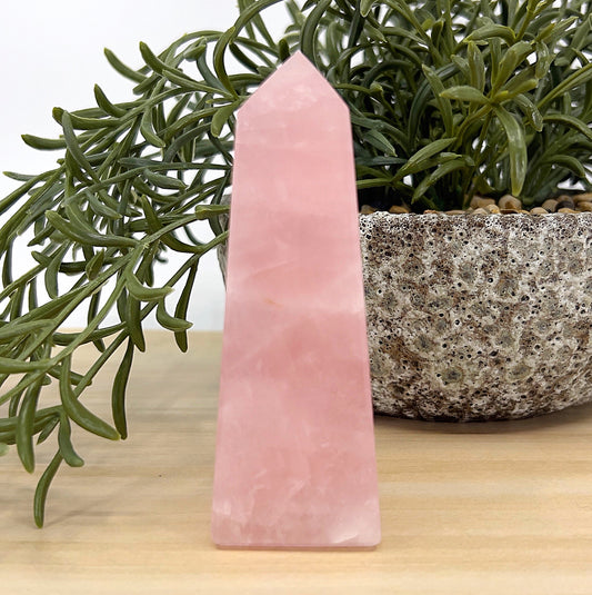 Rose Quartz obelisk crystal tower, 10cm tall, in soft pink tones beside a stone pot and greenery — symbolising love, emotional healing, and heart chakra balance.