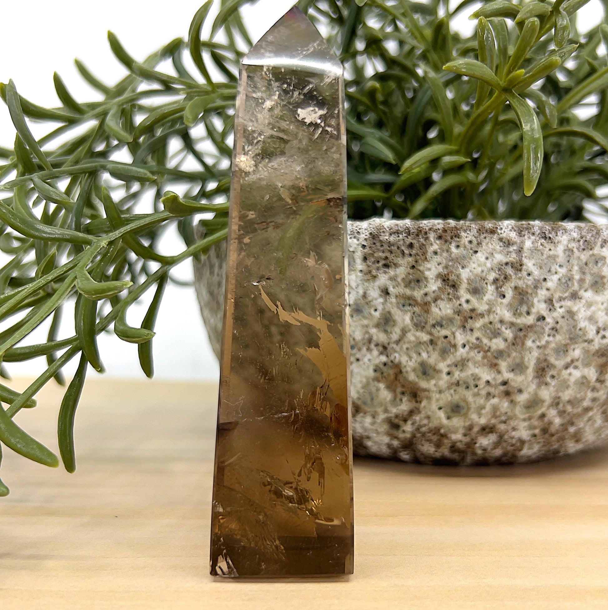 Smoky Quartz obelisk crystal tower 10 cm tall, translucent brown with natural inclusions, placed beside a stone pot and green foliage – symbolising grounding and protection.