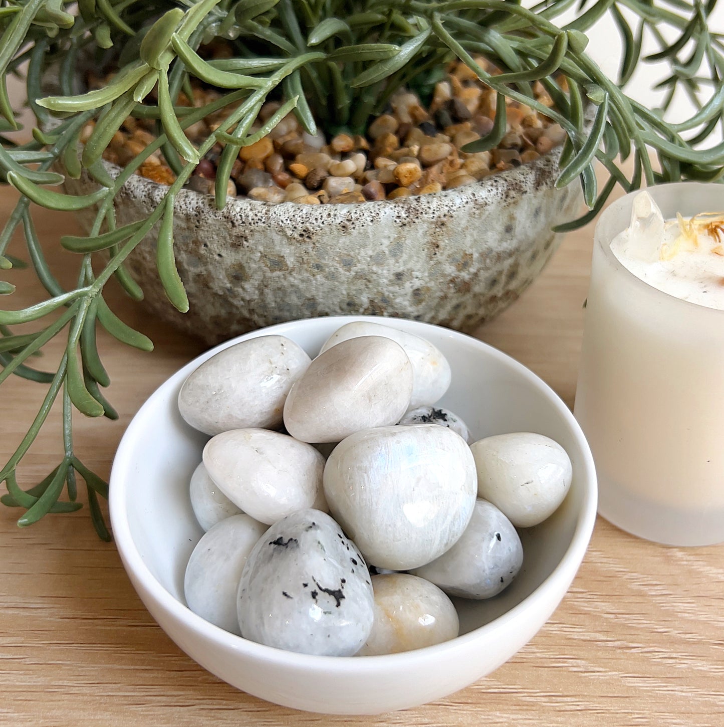White bowl, White Moonstone tumbles, potted plant, and candle on a wooden surface