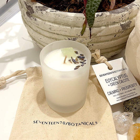 A vegan-friendly, soy wax candle by Seventeen70 Botanicals, infused with eucalyptus and green calcite, titled "Calming + Prosperity." The candle is set on a white background, adorned with dried lavender buds and a small piece of green calcite, emphasizing its natural and soothing properties. The candle is accompanied by a label and burlap pouch, enhancing its artisanal appeal.