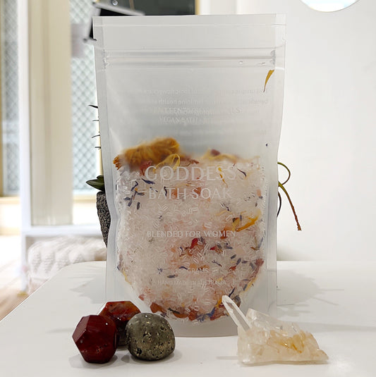A clear resealable pouch containing Seventeen70 Botanicals Goddess Bath Soak, 200g, displayed with vibrant dried flowers visible inside. The package is elegantly labeled with white text and is surrounded by crystals, including red jasper, pyrite, and clear quartz, placed on a white surface. In the background, a green potted plant adds a natural touch, enhancing the product's spa-like and holistic aesthetic. | Simply Esoteric
