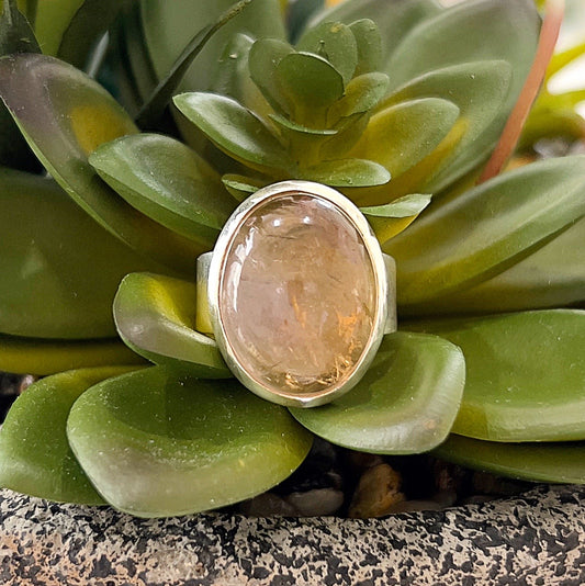 Close-up image of an ametrine sterling silver ring displayed against a backdrop of green succulents. The oval-shaped cabochon ametrine gemstone features a captivating blend of golden citrine and purple amethyst hues, set in a polished sterling silver bezel. The natural setting enhances the ring's earthy and elegant aesthetic. | Simply Esoteric