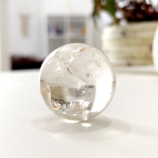 A perfectly polished clear quartz sphere, transparent and gleaming, showcasing natural inclusions and reflecting light beautifully. This sphere is a versatile tool for energy harmonization and spiritual clarity, ideal for meditation and decorative purposes. | Simply Esoteric