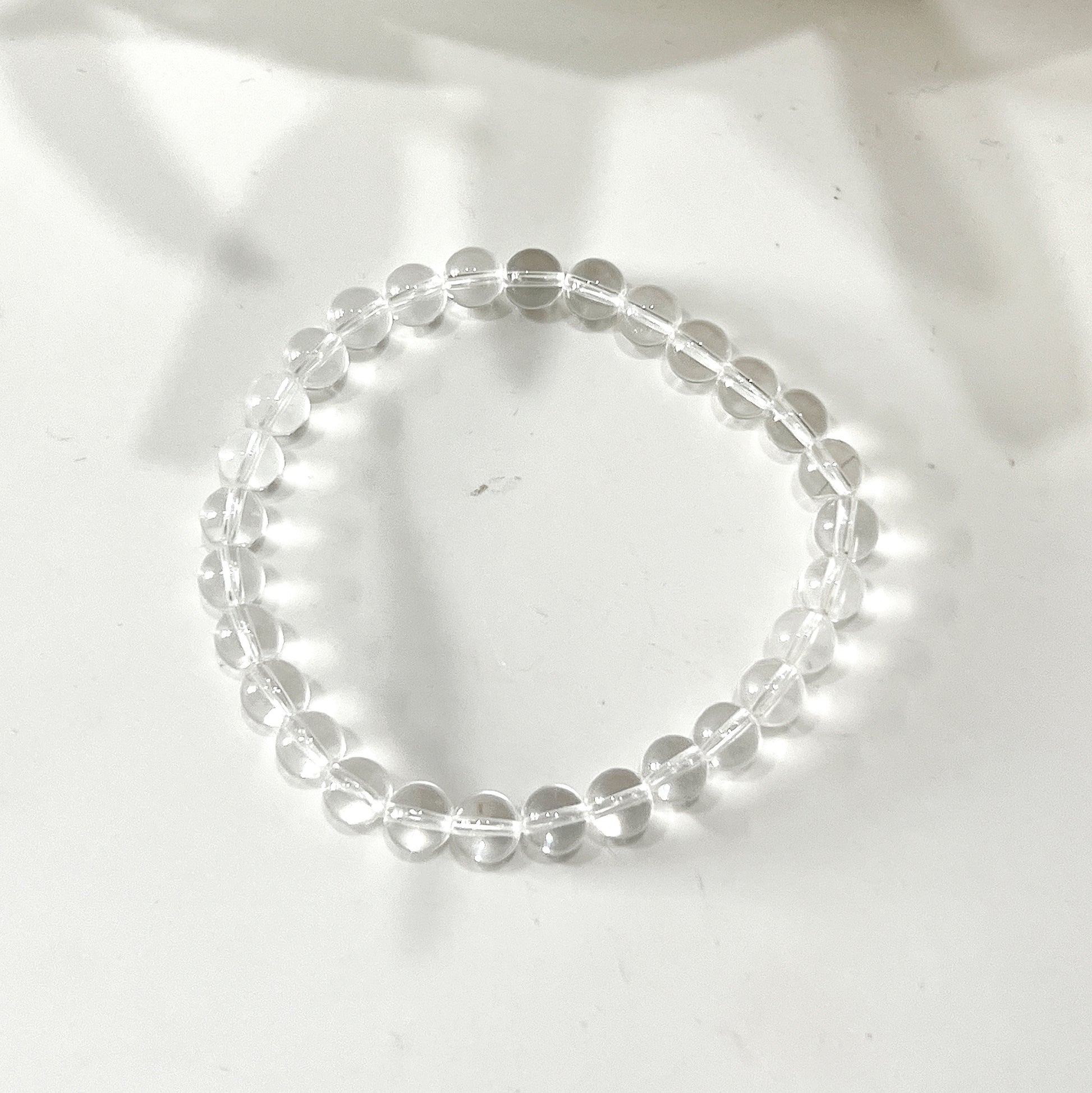 A clear quartz bead bracelet with 6mm round beads strung on a stretchy cord, arranged in a circle on a white background. The beads are transparent and evenly spaced, showcasing their natural clarity and shine. | Simply Esoteric