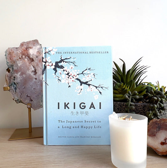 Light blue hardcover book titled *“Ikigai: The Japanese Secret to a Long and Happy Life”* by Héctor García and Francesc Miralles, standing on a wooden surface with a white candle, crystal cluster, and a potted succulent in the background. | Simply Esoteric