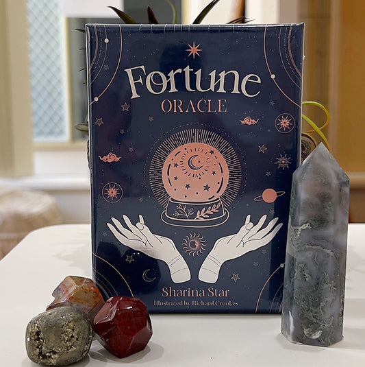 A neatly displayed set of *Fortune Oracle Cards* by Sharina Star, illustrated by Richard Crookes, featuring an elegant dark blue box with celestial-themed artwork, including a crystal ball, stars, planets, and mystical hands. The box is surrounded by polished gemstones and a crystal tower, placed in a serene setting with a light background, creating a mystical and inviting atmosphere. | Simply Esoteric