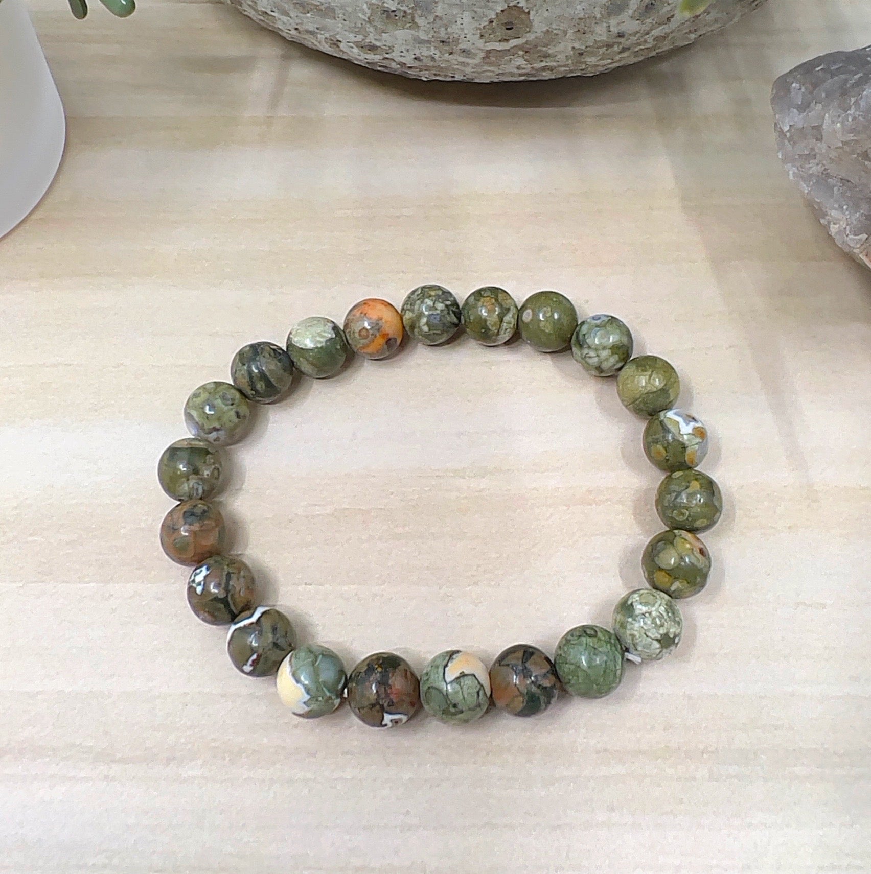 Handmade Rainforest Jasper crystal bead bracelet with 8mm round beads on stretchy cord, featuring earthy green, brown, cream, and orange tones, displayed on a wooden surface. | Simply Esoteric