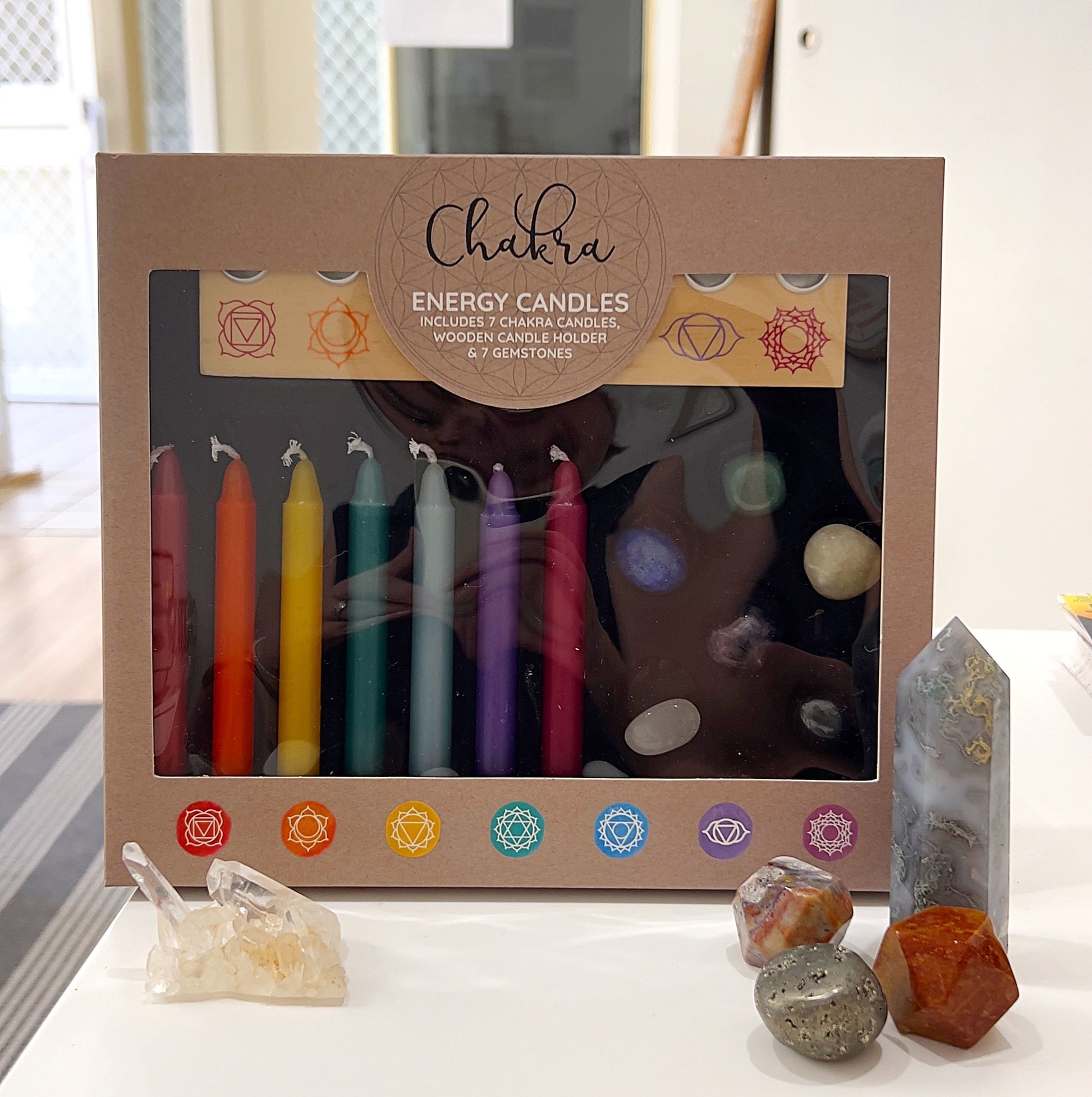 A Chakra Energy Candle and Crystal Gift Set displayed on a table. The set includes seven colored candles representing each chakra, a wooden candle holder, and seven chakra gemstones, all elegantly packaged in a decorative box. The background features natural elements, including crystal clusters and gemstones, enhancing the spiritual and calming aesthetic. | Simply Esoteric