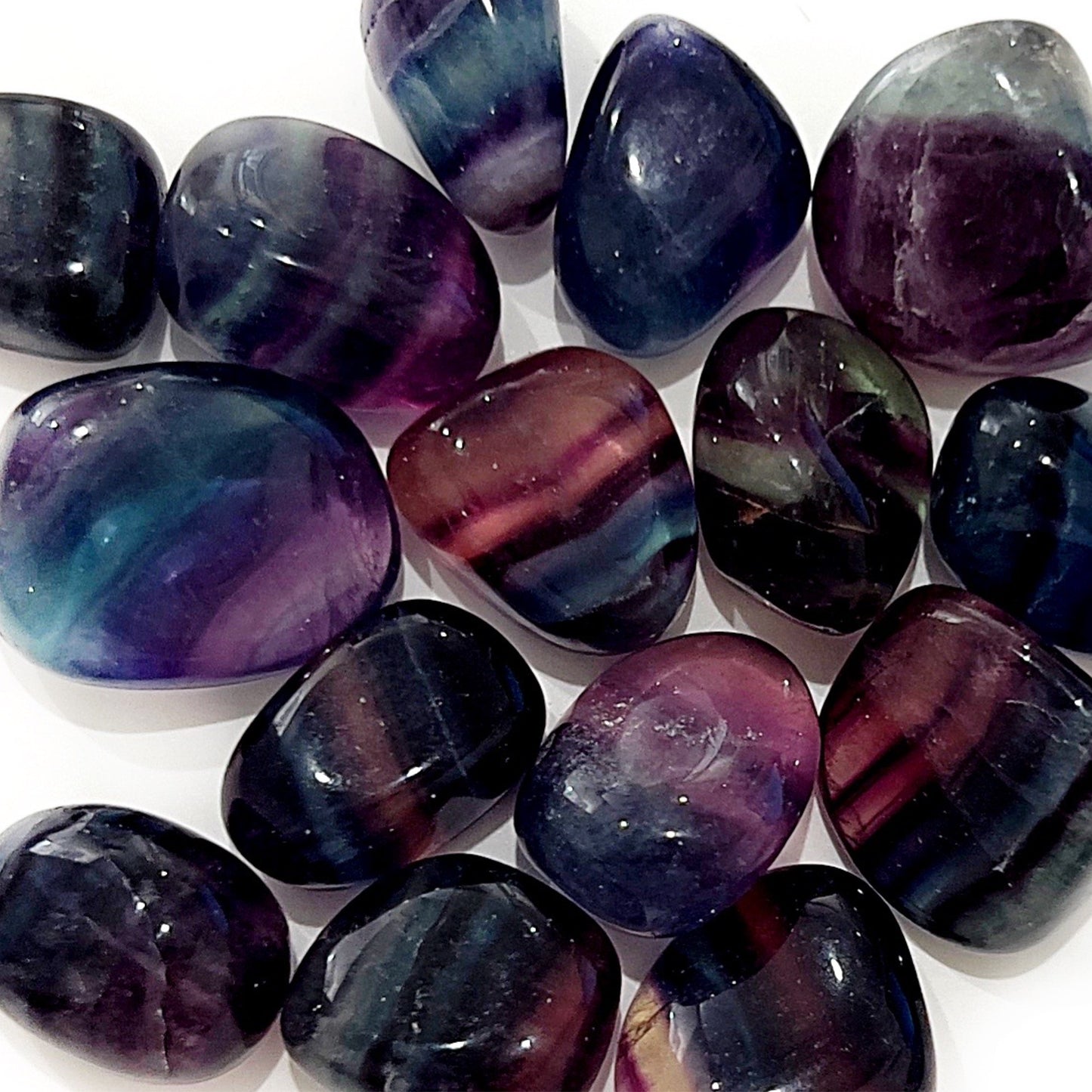 A vibrant assortment of Rainbow Fluorite tumbled stones, featuring a rich tapestry of deep purples, tranquil blues, and verdant greens. Each stone is polished to a lustrous sheen, with the varied translucency and banding unique to fluorite, all arrayed against a crisp white background. | Simply Esoteric