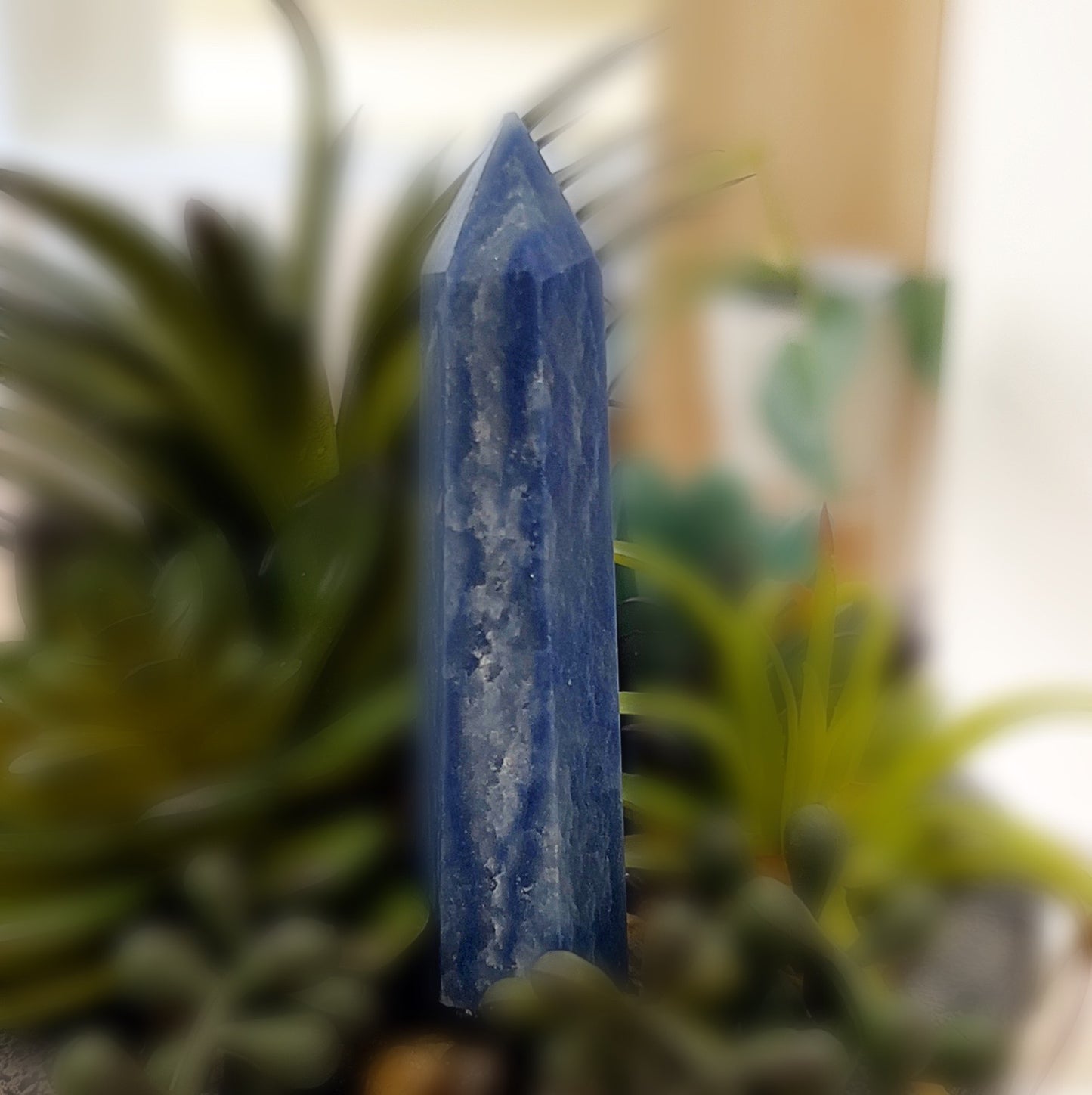 A tall, polished Blue Aventurine tower crystal, approximately 9cm in height, with a deep blue color and natural streaks. The tower is displayed against a soft, blurred background of green plants, emphasizing its smooth texture and calming presence. | Simply Esoteric