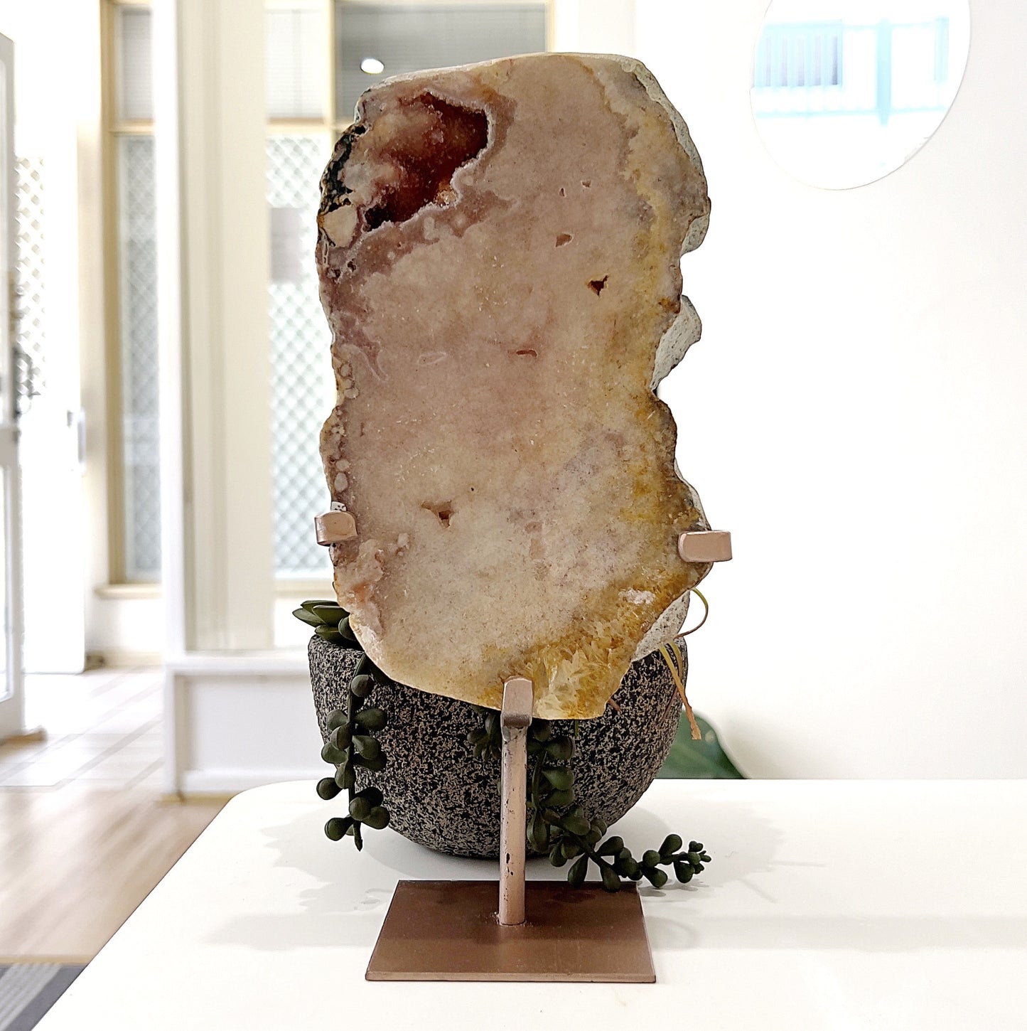 A large pink amethyst slice displayed on a rose gold-toned metal stand. The slice measures approximately 20cm tall and 11.5cm wide, showcasing natural soft pink and earthy tones with subtle crystalline formations. The total height, including the stand, is approximately 26.5cm. It is set against a backdrop of green succulents, highlighting its organic beauty. | Simply Esoteric