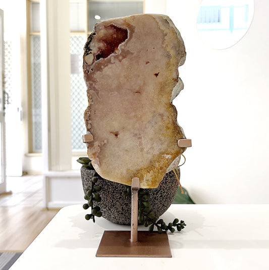 A large pink amethyst slice displayed on a rose gold-toned metal stand. The slice measures approximately 20cm tall and 11.5cm wide, showcasing natural soft pink and earthy tones with subtle crystalline formations. The total height, including the stand, is approximately 26.5cm. It is set against a backdrop of green succulents, highlighting its organic beauty. | Simply Esoteric