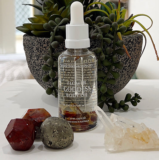 A clear glass bottle of Seventeen70 Botanicals Flower Goddess Body Oil, featuring a white dropper lid, placed against a natural backdrop of greenery in a decorative planter. The oil is light and infused with subtle botanicals, with small floral elements visible in the bottle. Surrounding the bottle are crystal accents, including a pyrite stone, red jasper, and a quartz cluster, enhancing the luxurious and natural aesthetic of the product. | Simply Esoteric