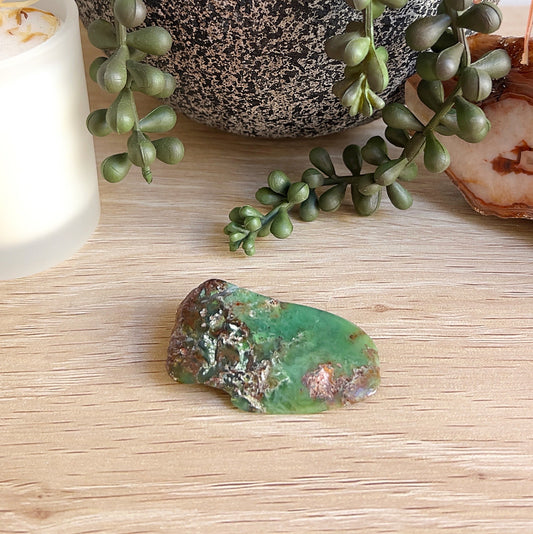 A polished piece of Western Australian chrysoprase, approximately 5cm in size and 19.8g, showcasing vibrant green tones with earthy brown and white mineral inclusions. Displayed on a wooden surface with a succulent plant, candle, and natural crystal slice in the background. | Simply Esoteric