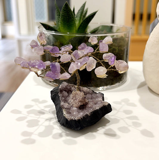 A handcrafted amethyst tree with polished amethyst crystals on delicate branches, rooted in a natural amethyst cluster base, displayed on a white surface with a green plant in the background.