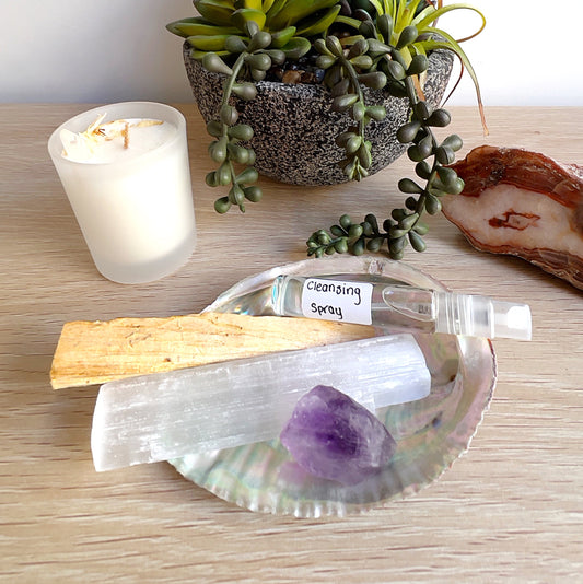 Home cleansing kit with palo santo stick, selenite wand, amethyst tumble, abalone shell, and cleansing spray displayed on a wooden surface with a candle and crystal decor in the background. | Simply Esoteric